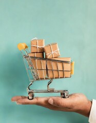 A person's hand holding a mini shopping cart filled with parcels against a turquoise background, representing online shopping and delivery 