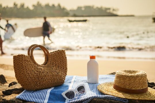 Beach scene with straw bag, sunscreen bottle, on blue towel. Sunglasses rest beside, essentials for sun safety, relaxation by sea. Sunny ocean backdrop with people, surfboard hint at active leisure.