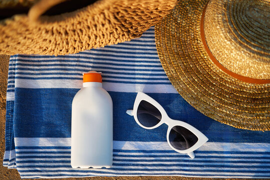 Overhead shot of summer essentials on sandy shore, white sunscreen bottle next to chic sunglasses, beside striped towel under woven sunhat for stylish sun protection at beach