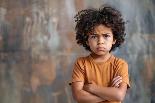 A young boy with voluminous curly hair stands scowling with his arms crossed
