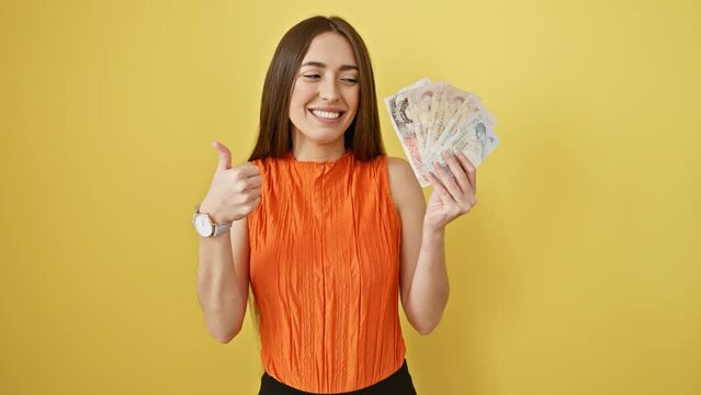 Joyous hispanic woman in studio, flashing an 'ok sign' and smiling brightly, holding english pounds banknotes on sunny yellow isolated background; portrait of confidence and business success