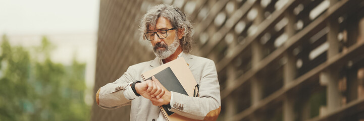 Panorama of mature businessman holding folder with business documents is standing outside on modern...