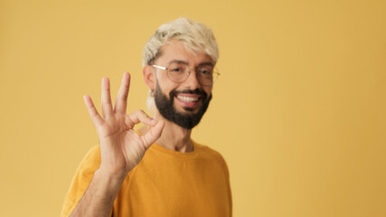 Guy with glasses, dressed in yellow T-shirt, showing ok gesture looking at camera isolated on yellow background in studio