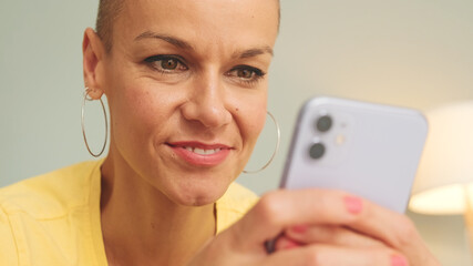 Close-up portrait of smiling bald woman relaxing on sofa in living room using mobile phone