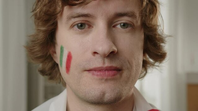 Man Italian football fan cheering for favorite Italy team, with painted flag on cheeks. Euro 2024 in Germany. World Cup 2026 USA, Canada, Mexico. Football, sports and soccer fans. Slow Motion