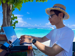 Nomad digital  young man with the laptop and running remotely with bright scenic view near poolside on the beach in summer time.