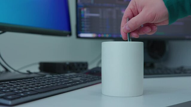A man's hand starts to stir a liquid with a spoon in a white mug that sits on a desk next to a computer monitor. The scene is likely a workspace. The coffee cup adds a sense of comfort