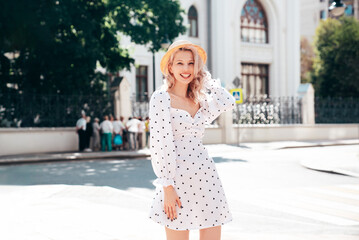 Young beautiful smiling blond woman in trendy summer white dress. Sexy carefree woman posing in street  at sunset. Positive model outdoors at sunny day. Cheerful and happy. In hat