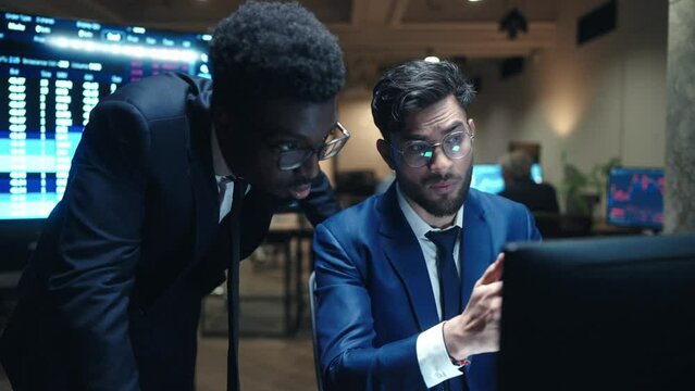 Two multi-ethnic men looking at screen in business office. Hardworking man in blue formal suit explaining something to director. Analysing stock market. On screen visible online data in real time.