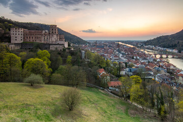 View over an old town with a castle or palace rune in the evening at sunset. This place is located in a river valley of the Neckar, surrounded by hills. Heidelberg, Baden-Württemberg, Germany - obrazy, fototapety, plakaty