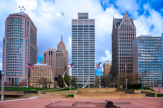 Detroit City vibrant skyline, high-rising buildings, modern plaza park with American Flags waving under dramatic white clouds in the blue sky in Michigan, USA