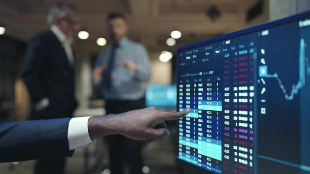 Camera view of computer display with financial data or crypto graphs. Man showing his fingers on display. Trading online. Stock market. Two men talking with each other in background. Business concept.