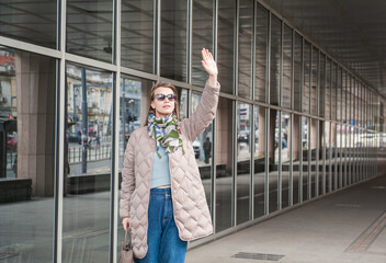 Beautiful stylish woman with sunglasses and bag walking near building with mirror wall on the street and waving her hand hello