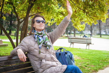 Beautiful stylish woman with sunglasses and bag sitting on the bench and waving her hand hello