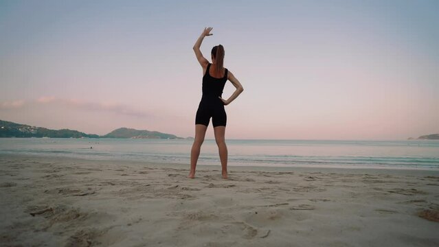 Barefooted woman doing yoga exercise lounge, warrior pose on sandy sea ocean beach at sunset. Female stretching legs. Body care, wellness, healthy lifestyle, morning sport outdoors training concept.