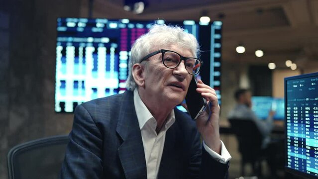 Caucasian broker talking with clients while using his phone. Intelligent man with business suit sitting at table and working at computer during remote communication. On screens visible graphs.