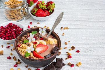 Chocolate smoothie bowl topped with oat granola, banana, strawberries, pomegranate and spring flowers on white wooden table. Healthy vegan protein food with cocoa for breakfast