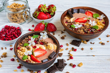 Chocolate smoothie bowl topped with oat granola, banana, strawberries, pomegranate and spring flowers on white wooden table. Healthy vegan protein food with cocoa for breakfast - 778878341
