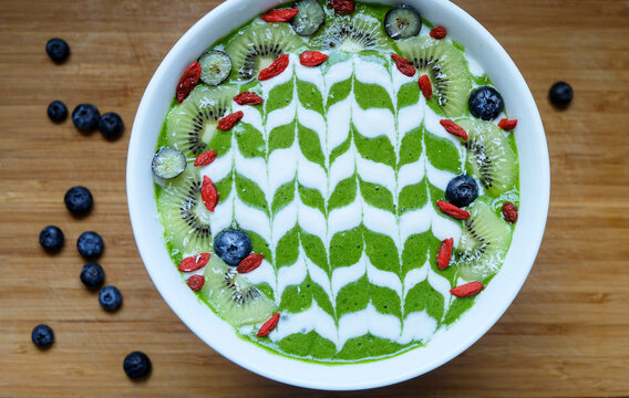 Green smoothie bowl made from kale, spinach, banana, pineapple and lime topped with fresh kiwi fruit, blueberry and goji berry with latte art deco style	