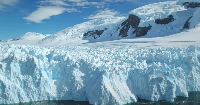 Huge towering ice glacier in Antarctica, snow covered mountain in background, clear blue sunny sky. Ice formation wall cold polar ocean. Aerial arctic winter landscape. Global warming, climate change