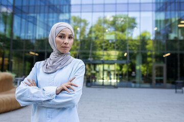 Professional Muslim woman with arms crossed in front of modern corporate architecture, exuding...