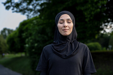 A young woman wearing a hijab stands outdoors with her eyes closed, exuding a sense of peace and serenity surrounded by nature.