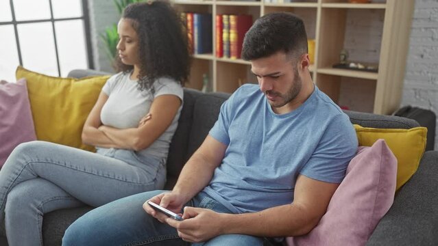 A distressed woman and an indifferent man sit separately with a phone on a sofa, suggesting relationship issues in a living room setting.