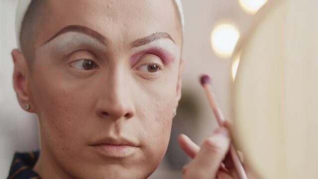 Young man in a drag queen makeup looking in a hand mirror and applying shadows to his eyes with a brush in the dressing room close up during preparation for the performance