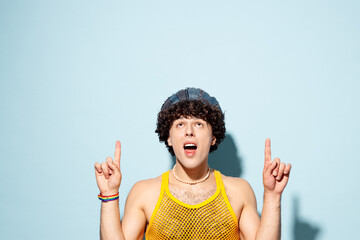 Young happy gay Latin man wear mesh tank top hat clothes point index finger overhead on empty area isolated on plain pastel blue cyan background studio portrait Pride day June month love LGBT concept