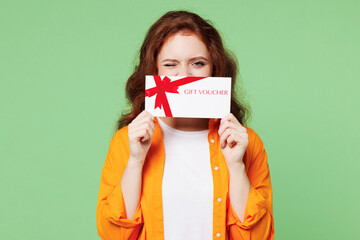 Young ginger woman she wear orange shirt white t-shirt casual clothes hold cover mouth with gift certificate coupon voucher card for store isolated on plain pastel green background. Lifestyle concept.