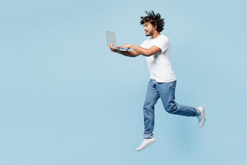 Full body side view young IT Indian man wear white t-shirt casual clothes jump high hold use work on laptop pc computer isolated on plain pastel light blue cyan background studio. Lifestyle concept