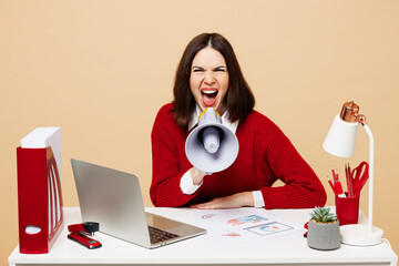 Young mad employee business woman wear red sweater sit work at office desk with pc laptop hold megaphone scream announces sale Hurry up isolated on plain beige background. Achievement career concept. - obrazy, fototapety, plakaty