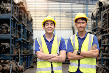 Portrait of two happy smiling Asian industrial engineer worker men with safety vest and helmet standing with arms crossed at manufacturing plant factory with many engine parts as blurred background.