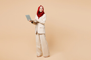 Full body fun young Arabian Asian Muslim IT woman wear red abaya hijab suit clothes hold use work on laptop pc computer look aside isolated on plain beige background. UAE middle eastern Islam concept.