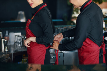 Smiling waitress asking coworker to tie apron - obrazy, fototapety, plakaty