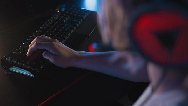 Left-handed gamer plays video game at computer club Cyber sport tournament. E-sport championship. Fingers press keys on gaming RGB keyboard, rack focus to headset. Skill. Multiplayer strategy shooter