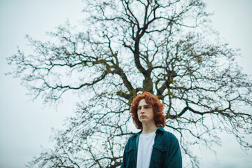 Young man with red hair standing under a bare tree, gazing into the distance