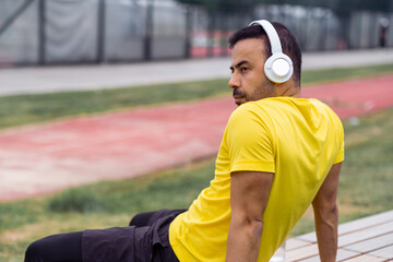 man athlete, wearing wireless headphones resting on wooden bench, taking a break after a workout at...