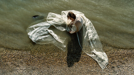 young nude sexy mermaid woman sitting in a transparent white cloth dress on the bank of the river Danube wet in shallow water