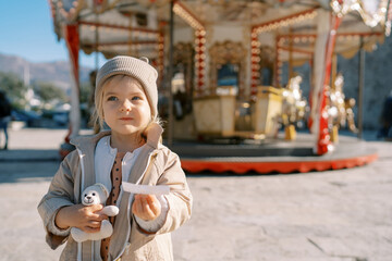 Little girl with a teddy bear stands with a ticket in her hand near a spinning carousel