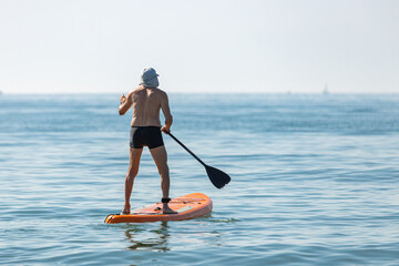 Senior man using a sup board on a calm, blue sea on a hot summer day during a heatwave; old man standing on a paddleboard on the ocean.