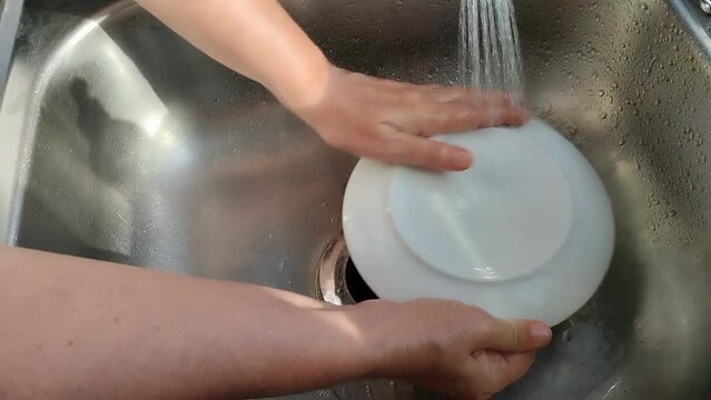 Unas manos de mujer lavando  un plato de color blanco con  agua en un fregadero