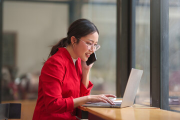 Fototapeta na wymiar Asian businesswoman in a red jacket is talking on her cell phone while using a laptop