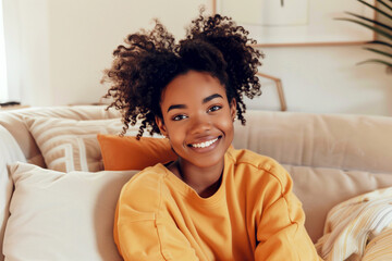 A young African-American smiling girl in an orange sweatshirt sits and relaxes on the sofa. Modern minimalist interior design in beige pastel colors