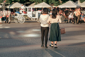 Two close Gen Z lesbian friends walk arm in arm towards a white carriage in an old square, gender...