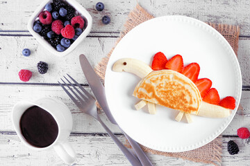 Fun child theme breakfast pancake in the shape of a dinosaur. Top view table scene on a white wood background. - 779043363