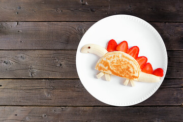 Cute child theme breakfast pancake in the shape of a dinosaur. Top down view on a dark wood table background.