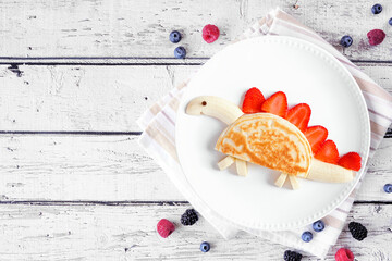 Cute child theme breakfast pancake in the shape of a dinosaur. Above view table scene on a white wood background. Copy space.