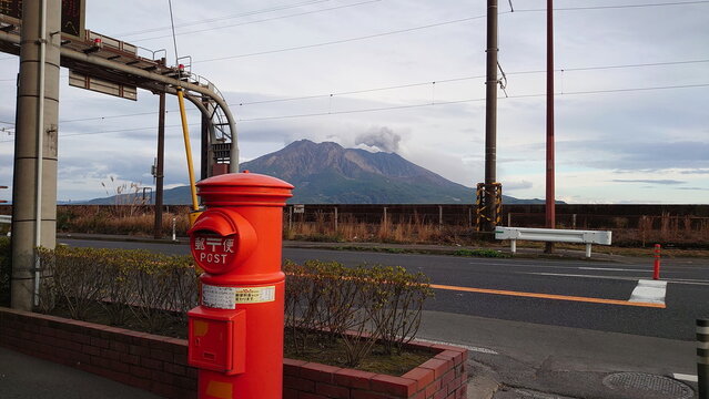 Kagoshima, Japan - 01.29.2020: Sakurajima releases smoke under a blue sky with a red Japanese postbox in the front on the pavement along an empty road