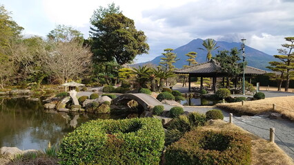 Kagoshima, Japan - 01.29.2020: A stone bridge over a pond with a pavilion on the side in Sengan-en and Sakurajima at the back releasing smoke under a cloudy sky